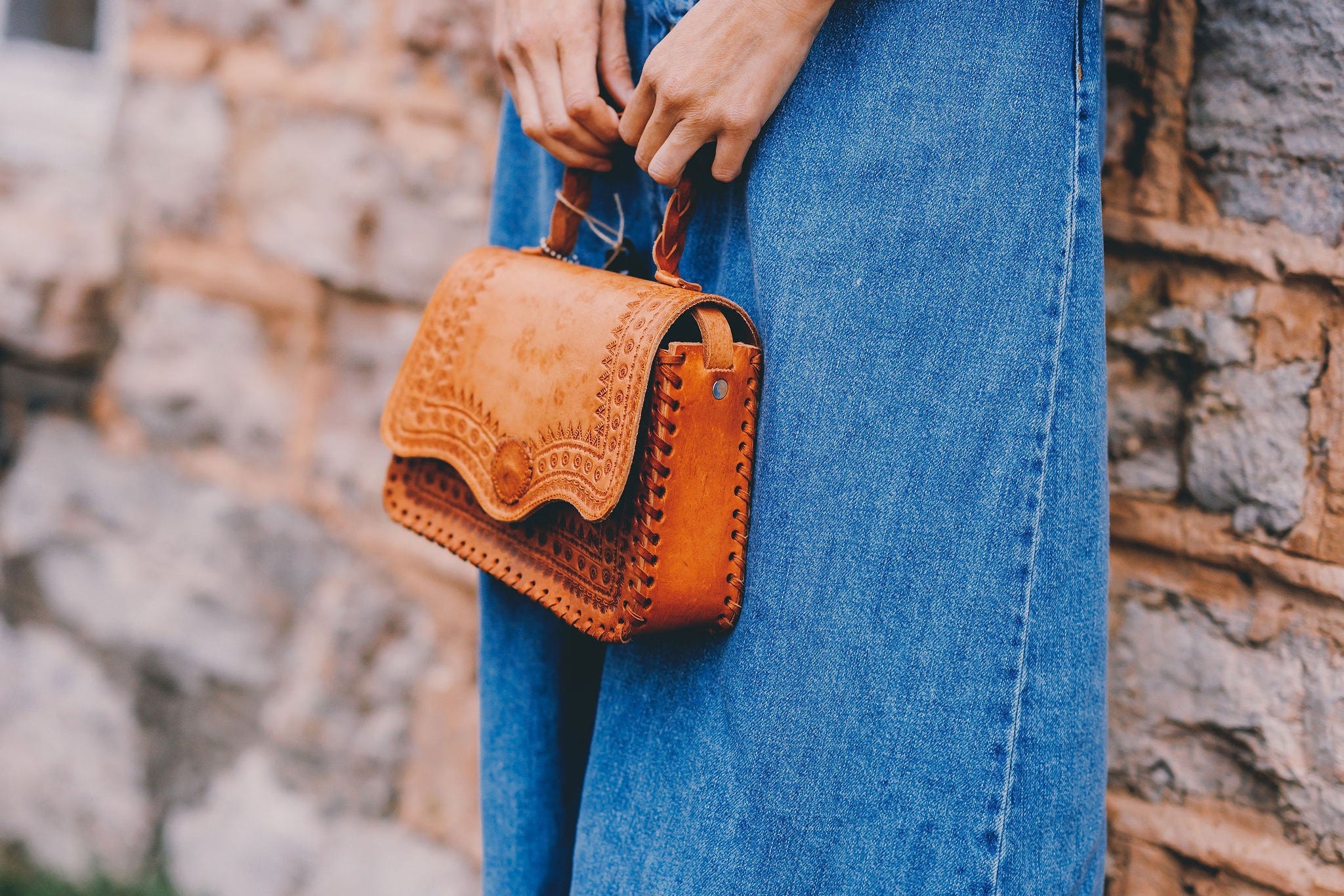 Person wearing a long denim dress holding a brown leather handbag against a stone wall.