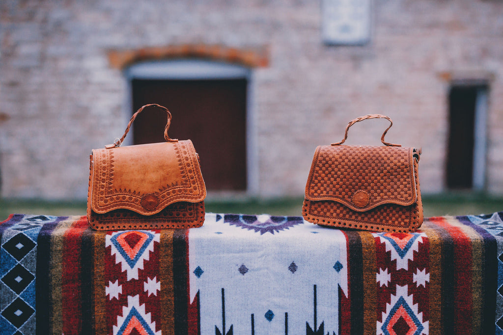 Two brown leather handbags on a colorful patterned surface with a stone building in the background.