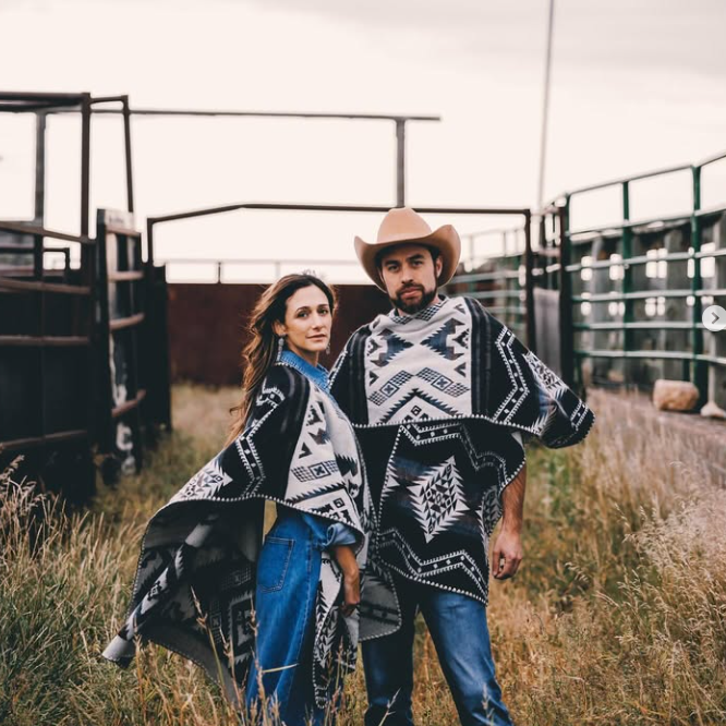 Two people wearing patterned ponchos standing in a grassy field with a fence in the background.